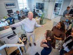 Students gather around a man who is talking and pointing, with people working at their desks in the background.