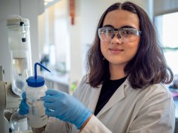 Student wearing safety glasses and gloves holds a lab cylinder.