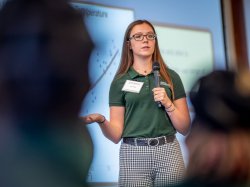 A female student dressed in green gestures as she speaks into a microphone.