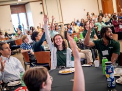 People raise their hands while seated at conference room tables.