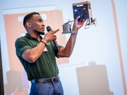 A student holds up a model sensor while speaking onstage.