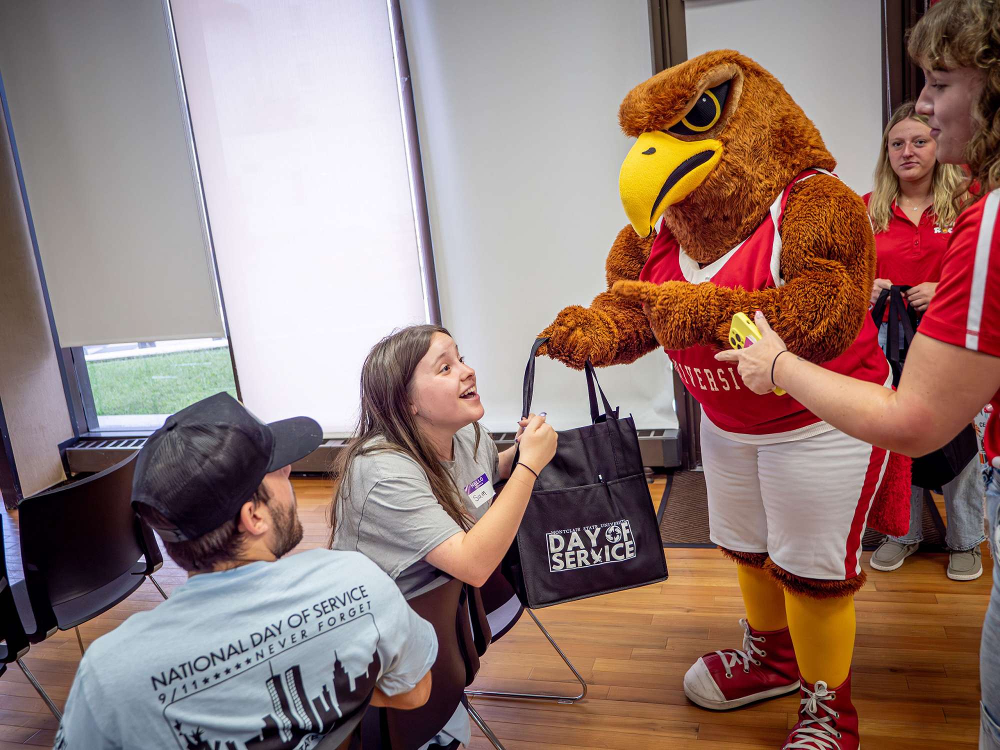A college mascot, a giant red hawk, gives a student a black bag with the words “Day of Service” on it.