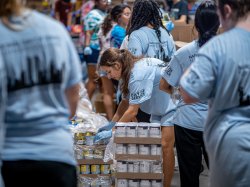 A student bends down to unpack cans in a crowded warehouse.