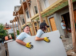 Two people carry a large piece of drywall material at a construction site.