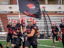 The Red Hawks football team runs onto the field carrying a black and red Red Hawks Football flag.