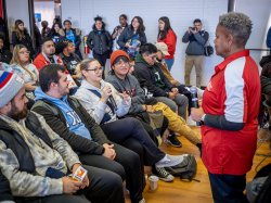 A group of seated students talk with a woman standing in red and white.