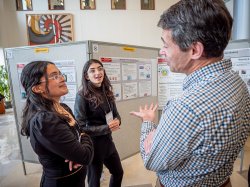 Two young scientists talk to a professor in front of a research poster.