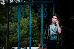 A young person stands at a fence, next to a guitar, talking on the phone.
