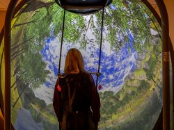 A student wearing a harness stands in the Bertec balance system with a virtual outdoor scene before her.