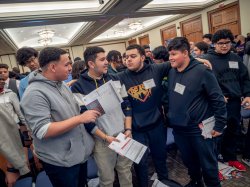 A male student put his arms around classmates in a roomful of young male high school students.