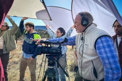 A man with earphones and a camera points as other people hold lighting equipment overhead.