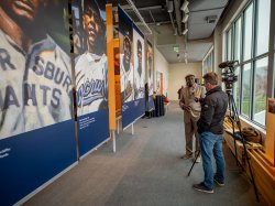 A man with a camera and a microphone interviews another man in front of a line of banners featuring players.