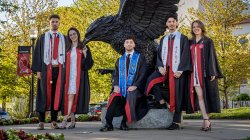 The Povolo quintuplets in graduation gowns stand in front of a large Red Hawk statue.