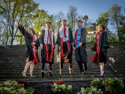 The Povolo quintuplets wearing graduation gowns jump together in front of stone steps outdoors.