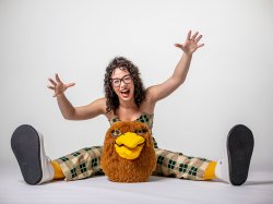 Eliza Andrus laughs and opens her arms wide sitting behind Ricky’s mascot head and wearing oversized sneakers.