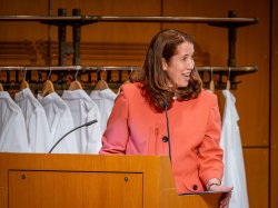 Amy Aiello speaks at a lectern before a rack of white lab coats.