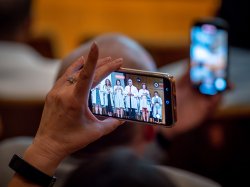 A hand holds a phone showing Audiology students on stage.