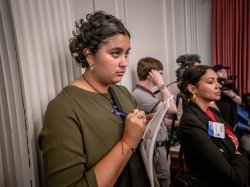 A woman standing in the back takes notes.
