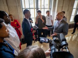 A student and legislator talk in the hallway surrounded by people and a camera with a microphone.