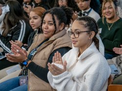 A group of high school students sitting in an audience clap their hands during the Women’s Leadership Conference at 火狐体育. The foreground shows two women, one wearing a beige vest and another wearing glasses and a white fleece, both engaged and smiling. The background reveals more attendees focused on the event.