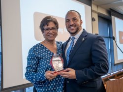 Carmen Reyes Cuevas, wearing a blue patterned blouse, holds an award as she poses with EOF alumnus Rey Acevedo, who is dressed in a suit and tie. Both are smiling in front of a presentation screen at the event.