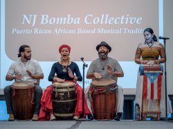 A group of four performers from the NJ Bomba Collective playing traditional drums on stage, with one performer singing and another speaking at a podium adorned with the Puerto Rican flag. A screen behind them displays "NJ Bomba Collective: Puerto Rican African Based Musical Tradition."