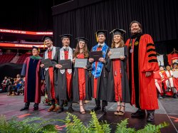 The Povolo五胞胎, wearing graduation caps and gowns, stand together on stage at 火狐体育's 2024 Commencement ceremony. Each holds their diploma while smiling alongside university officials in academic regalia.