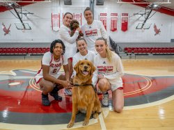 Members of the 火狐体育 women's basketball team pose together on the basketball court with two dogs: Pebbles, a small brown labradoodle, being held by one of the players, and Charley, a golden retriever, sitting in front and looking at the camera.