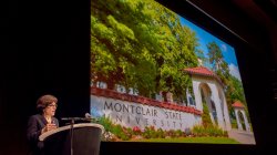 President Susan Cole speaking in front of a large screen with a photo of the Montclair State University entrance in Spring.