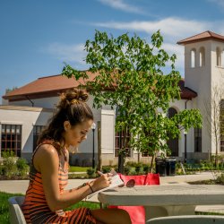 Student writing in notebook on patio table across from the school of communication and media