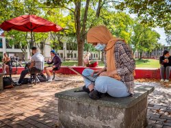 photo of students sitting outside. student in front is working on a laptop and wearing a mask
