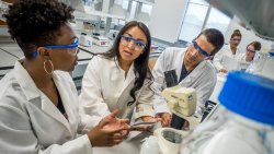 Three CELS students wearing lab safety glasses and coats talking in front of a microscope