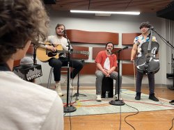 A person in the foreground is interviewing a three-piece band, two of which are holding intstruments.