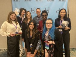 Eight people standing and smiling in front of a branded backdrop. Six of them are students who were recognized for winning national radio production awards.