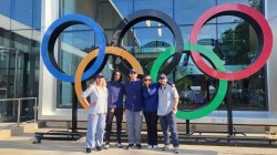 Five people standing and smiling in front of the Olympic Rings, which are large, colored circles.