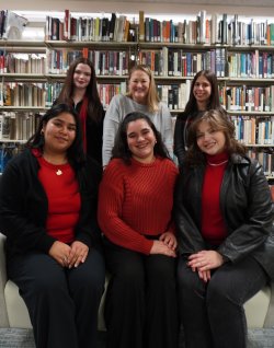 A group of five students and their professor. Three students are sitting in the front row and three people are standing in the back. They are in a library with a bookshelf behind them. They are smiling.