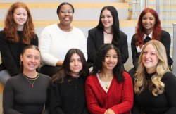 A group of eight students, sitting in two rows in equal groups of four. They are dressed professionally and smiling