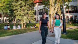 Two female students walking along a campus path with other students sitting on the lawn at Montclair State University.