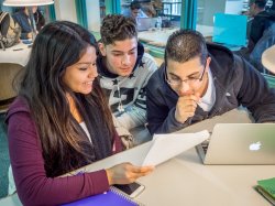 Photo of three students writing and collaborating in the library with a paper and a laptop.